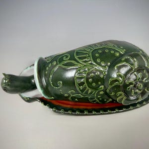 May include: A dark green ceramic decorative container with a lid. The container has a spiral design on the side and a small handle. The lid is a matching dark green color with a spiral design. The container sits on a red and green base.