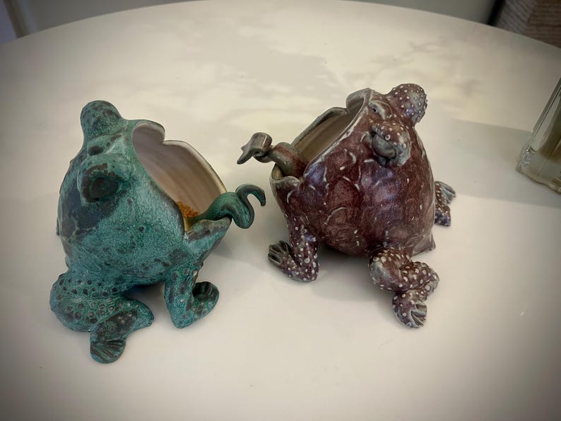Puede incluir: Dos macetas de cer&aacute;mica con forma de rana, una verde y otra marr&oacute;n, con la boca abierta y detalles elaborados. La maceta de rana verde tiene un interior marr&oacute;n y la maceta de rana marr&oacute;n tiene un interior blanco.