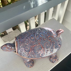 May include: A ceramic turtle figurine featuring a mottled blue and grey glaze with raised copper-colored swirling patterns across its shell, head, and legs. The turtle has a distinct head extending from the shell and four textured feet.