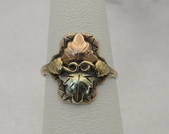 May include: A gold ring featuring a three-toned leaf design. The leaf is surrounded by a gold frame and has a vine design on either side.
