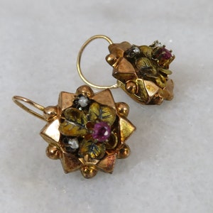 Peut inclure: Une paire de boucles d'oreilles en or avec un motif en étoile. Les boucles d'oreilles sont décorées de petites feuilles vertes, de pierres rouges et blanches et de perles dorées.