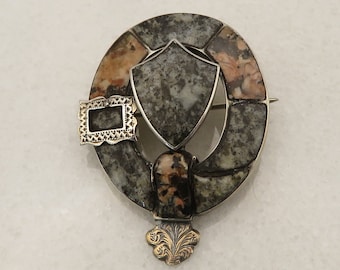 May include: A silver and gold brooch with a circular design featuring a shield and a buckle. The brooch is made of metal and has a stone inlay.