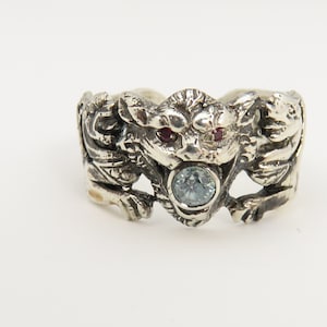 May include: A silver ring with a detailed design of a mythical creature with red eyes and a blue gemstone in the center.