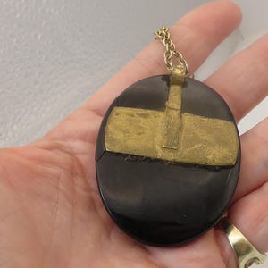 May include: A black oval pendant with a gold-colored metal overlay in a cross-like design. The pendant is attached to a gold-colored chain. The jewelry is held in a hand.