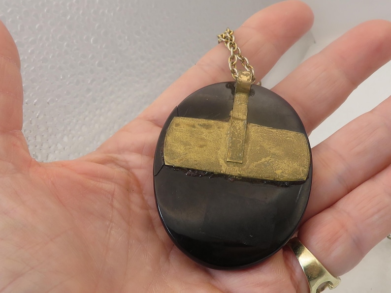 May include: A black oval pendant with a gold-colored metal overlay in a cross-like design. The pendant is attached to a gold-colored chain. The jewelry is held in a hand.