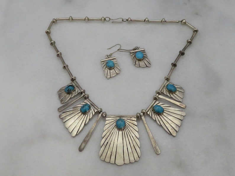 May include: A silver necklace and earrings set with turquoise stones. The necklace features a geometric design with multiple fan-shaped pendants. The earrings are simple and feature a single turquoise stone.
