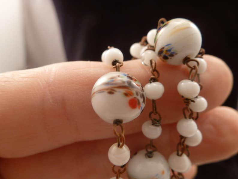 May include: A white glass beaded necklace with a brown wire chain. The beads are decorated with colorful floral patterns.