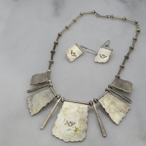 May include: A silver necklace and earrings set with a geometric design. The necklace has a chain with multiple pendant shapes. The earrings are dangle style with a similar geometric design. The back of the necklace and earrings are stamped with the number "925", indicating sterling silver.