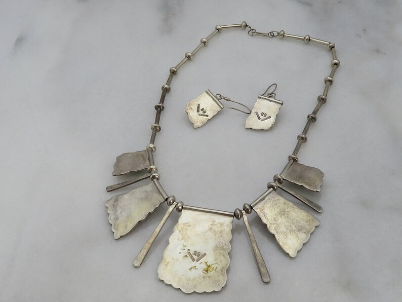May include: A silver necklace and earrings set with a geometric design. The necklace has a chain with multiple pendant shapes. The earrings are dangle style with a similar geometric design. The back of the necklace and earrings are stamped with the number "925", indicating sterling silver.