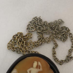 May include: A vintage cameo pendant necklace featuring a detailed relief carving of a classical figure. The pendant is set in a black frame and hangs from a silver-tone chain. The cameo has a light brown background with white figures.