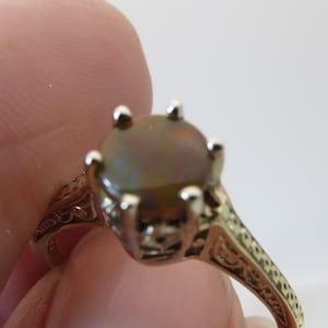 May include: A gold-toned ring with an oval, iridescent gemstone. The ring features an ornate, filigree design on the band and around the gemstone setting. The gemstone has a mix of brown, green, and gold hues.