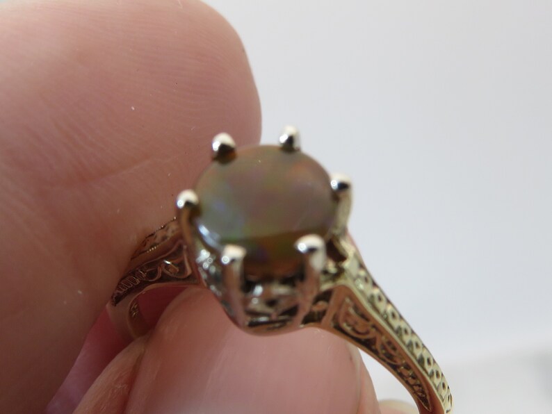 May include: A gold-toned ring with an oval, iridescent gemstone. The ring features an ornate, filigree design on the band and around the gemstone setting. The gemstone has a mix of brown, green, and gold hues.