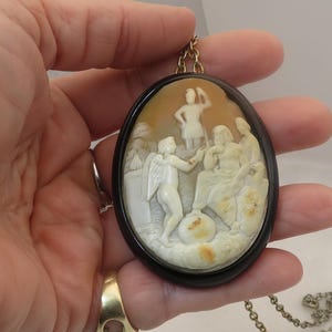 May include: Oval cameo pendant with a detailed relief carving of classical figures in cream and tan tones, set in a dark brown frame. The pendant hangs from a gold-tone chain, showcasing a piece of classical art.