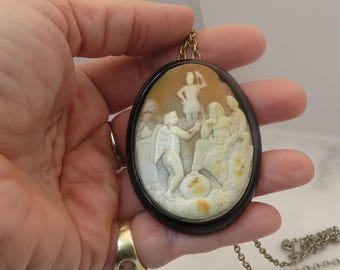May include: Oval cameo pendant with a detailed relief carving of classical figures in cream and tan tones, set in a dark brown frame. The pendant hangs from a gold-tone chain, showcasing a piece of classical art.