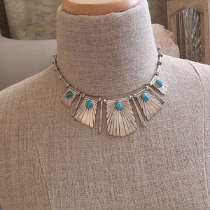 May include: A silver necklace with turquoise stones and a geometric design. The necklace is on a mannequin.