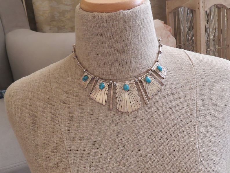 May include: A silver necklace with turquoise stones and a geometric design. The necklace is on a mannequin.