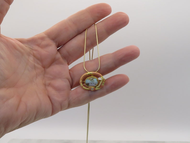 May include: A gold necklace with a pendant featuring a pink and gold oval-shaped stone surrounded by a gold snake design.