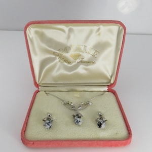 May include: A pink velvet jewelry box with a white satin interior. The box contains a silver necklace and earrings with a white and black stone pendant. The box has the text "Orobelle Valley of Treasure" on the lid.