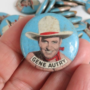 May include: A vintage blue and white button featuring a portrait of Gene Autry, a famous American singer, actor, and cowboy. The button has a red and white cowboy hat and a red bandana. The text "GENE AUTRY" is printed on the button.