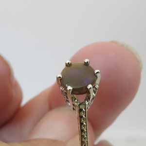 May include: Close-up of a ring with a dark, oval-shaped gemstone. The ring has a silver-colored band with intricate filigree detailing. The gemstone is set in a four-prong setting, and the ring is held by a person's finger.
