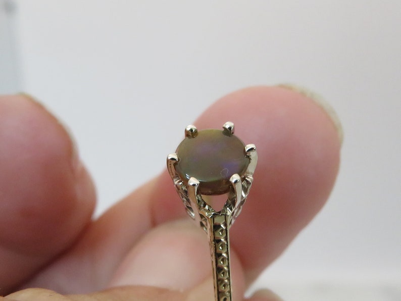 May include: Close-up of a ring with a dark, oval-shaped gemstone. The ring has a silver-colored band with intricate filigree detailing. The gemstone is set in a four-prong setting, and the ring is held by a person's finger.