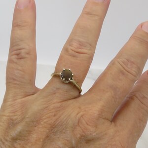 May include: A gold ring with a round, dark brown gemstone. The ring's band has a textured, twisted design and is set against a neutral background. The ring is on a person's finger.