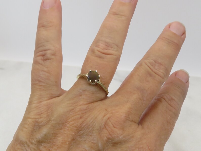 May include: A gold ring with a round, dark brown gemstone. The ring's band has a textured, twisted design and is set against a neutral background. The ring is on a person's finger.