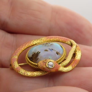 May include: A gold snake brooch with an opal and diamond accents. The snake is coiled around a large oval opal with a gold setting. The snake's body is textured and has a pink enamel finish.