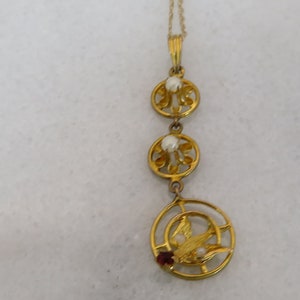 May include: A gold necklace with a pendant featuring three circular designs with small white pearls and a central design with a gold bird and a red gemstone.