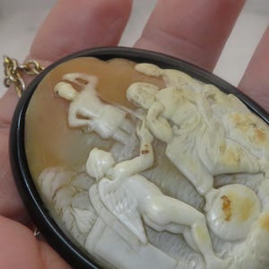 May include: Oval cameo pendant with a black frame and gold chain. The cameo features a detailed relief carving of classical figures in cream and tan tones. The figures are in a dynamic pose, suggesting movement and action.