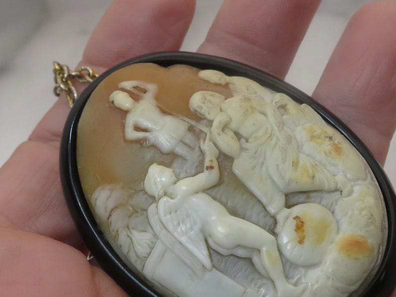 May include: Oval cameo pendant with a black frame and gold chain. The cameo features a detailed relief carving of classical figures in cream and tan tones. The figures are in a dynamic pose, suggesting movement and action.