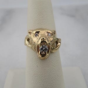May include: A gold-colored ring featuring a bear's head design. The bear's eyes and mouth are accented with small, sparkling gemstones. The ring is displayed on a white finger form.