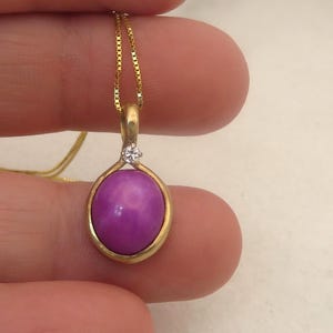May include: A gold pendant necklace with a vibrant purple gemstone. The pendant has a gold bezel and a small, clear accent stone. The chain is a delicate gold colour. The gemstone is approximately 1.5 cm in diameter.