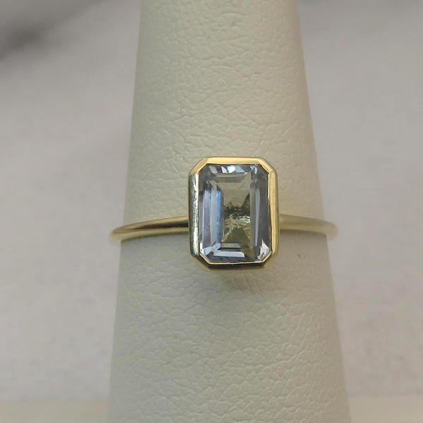 May include: A gold ring featuring a rectangular, light blue gemstone. The gemstone is set in a bezel setting, and the band is a thin, polished gold. The ring is displayed on a white surface.