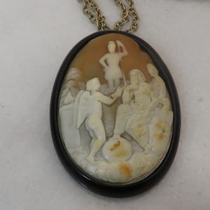 May include: Oval cameo pendant with a detailed relief carving of classical figures in cream and tan tones, set in a black frame. The pendant hangs from a gold-colored chain, showcasing a vintage or antique aesthetic. The carving depicts multiple figures in a scene.