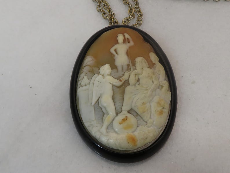 May include: Oval cameo pendant with a detailed relief carving of classical figures in cream and tan tones, set in a black frame. The pendant hangs from a gold-colored chain, showcasing a vintage or antique aesthetic. The carving depicts multiple figures in a scene.
