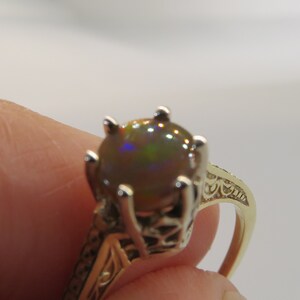 May include: A gold ring featuring a large, round, iridescent gemstone. The gemstone displays a play of colors, including green, blue, and brown. The ring has an ornate, filigree design on the band.