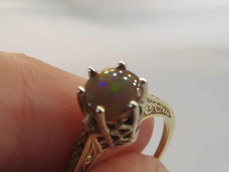 May include: A gold ring featuring a large, round, iridescent gemstone. The gemstone displays a play of colors, including green, blue, and brown. The ring has an ornate, filigree design on the band.