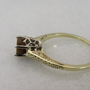 May include: A gold ring with an ornate band and a square, brown gemstone. The setting is detailed with filigree, and the ring is set against a neutral background. The ring is a classic piece of jewelry.