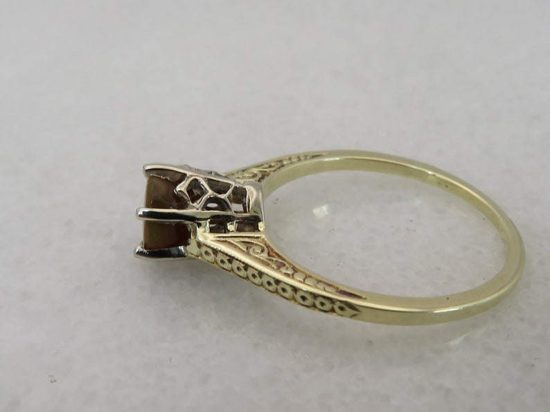 May include: A gold ring with an ornate band and a square, brown gemstone. The setting is detailed with filigree, and the ring is set against a neutral background. The ring is a classic piece of jewelry.