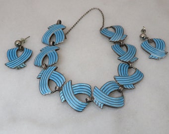 Puede incluir: Un collar y pendientes de plata con esmalte azul. El collar está hecho de piezas de esmalte azul curvadas y unidas. Los pendientes son simples tachuelas con esmalte azul.
