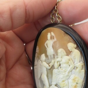 May include: Oval cameo pendant with a black border and gold-colored chain. The cameo features a detailed relief carving of classical figures in cream and beige tones. The pendant is held by a hand.