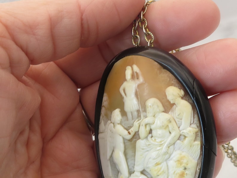 May include: Oval cameo pendant with a black border and gold-colored chain. The cameo features a detailed relief carving of classical figures in cream and beige tones. The pendant is held by a hand.