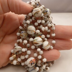 May include: A white and gold necklace with white glass beads and colorful painted details. The necklace is made of a chain with small white beads and larger white beads with colorful painted designs.