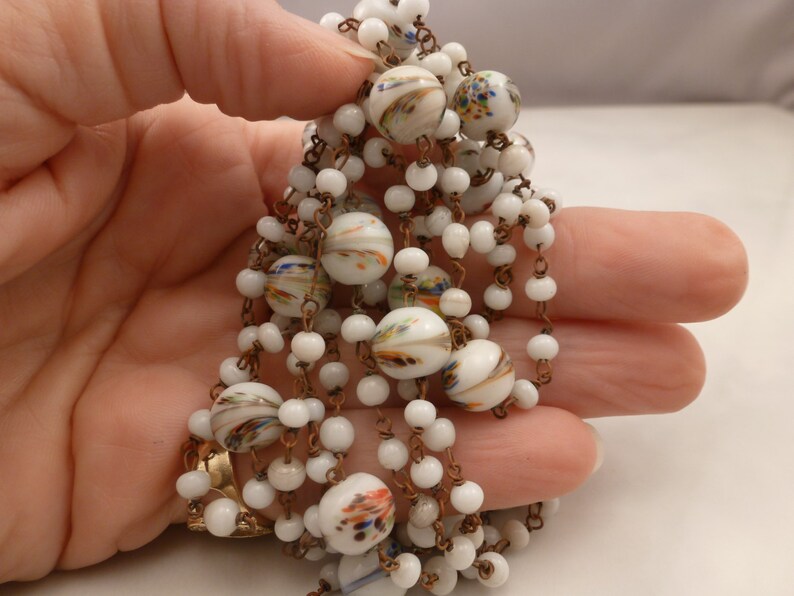 May include: A white and gold necklace with white glass beads and colorful painted details. The necklace is made of a chain with small white beads and larger white beads with colorful painted designs.