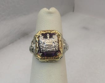 May include: A two-tone ring with a rectangular, deep purple stone at its center. The ring features a gold-colored frame and a silver-colored crest design. The crest includes a crown and other ornate details.