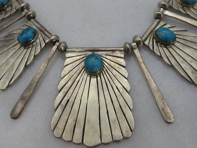 May include: A silver necklace with turquoise stones and a fan-shaped design. The necklace is made of sterling silver and features a unique geometric pattern.