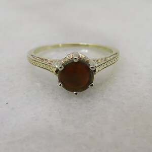 May include: A gold-toned ring with an oval, dark brown gemstone. The ring features a detailed band with an engraved pattern. The gemstone is held in place by six prongs. This piece of jewelry is suitable for everyday wear or special occasions.