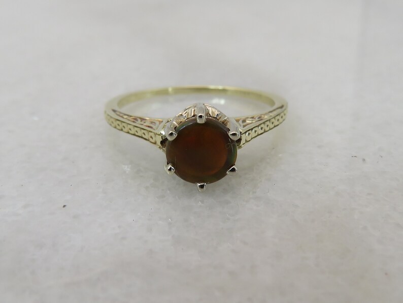 May include: A gold-toned ring with an oval, dark brown gemstone. The ring features a detailed band with an engraved pattern. The gemstone is held in place by six prongs. This piece of jewelry is suitable for everyday wear or special occasions.