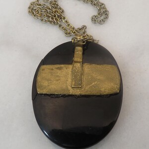 May include: A vintage necklace featuring a large, oval black pendant with a gold-colored metal overlay. The pendant hangs from a silver-colored chain. The gold metal has a rectangular design.